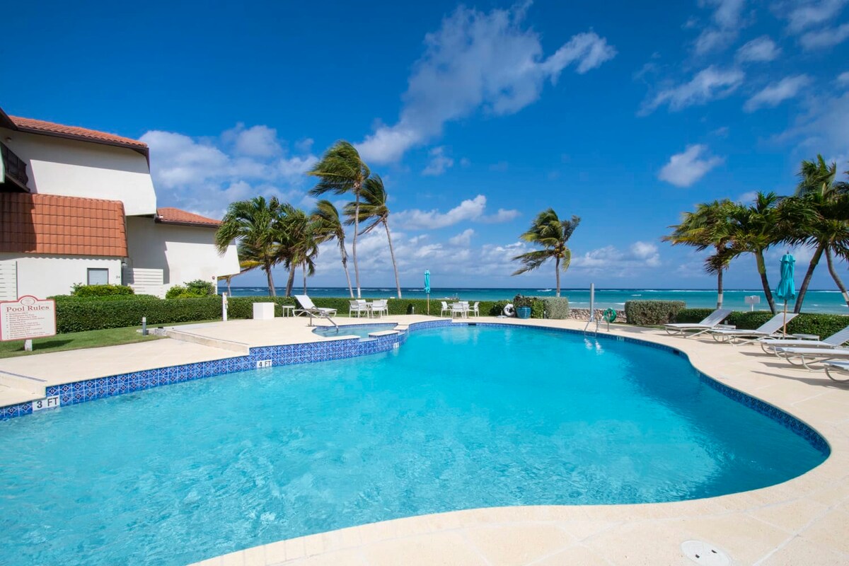 A refreshing swimming pool is set against a backdrop of palm trees and the Caribbean Sea. Sun loungers are positioned around the pool area, offering a relaxing space to enjoy the outdoors. The clear blue water reflects the bright sky, enhancing the serene atmosphere.