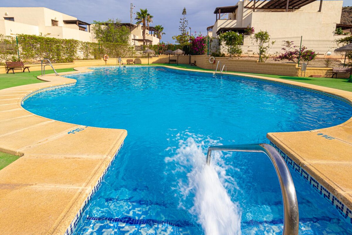 A large community pool is showcased, featuring clear blue water and a gentle splash from a water fountain. Sun loungers and lush greenery line the pool area, with residential structures visible in the background, creating a serene atmosphere for relaxation.