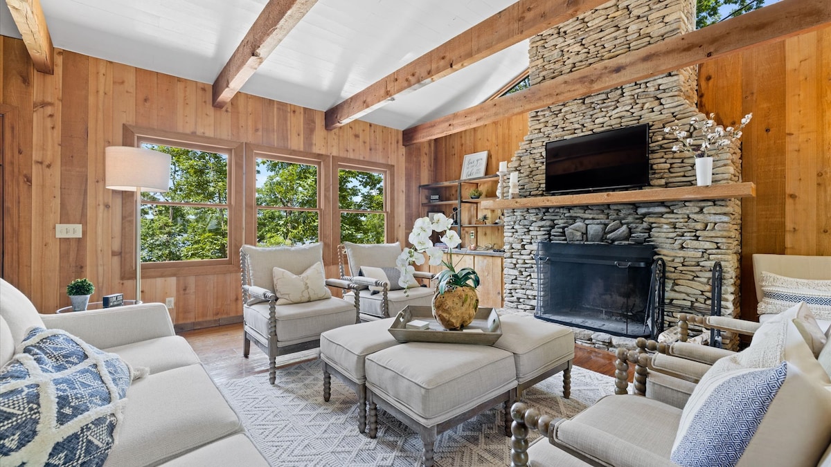 A cozy living area is presented with wooden walls and a stone fireplace. Light beige sofas and armchairs are arranged around a central coffee table, featuring decorative elements like a plant and a bowl. Large windows allow natural light to illuminate the space.