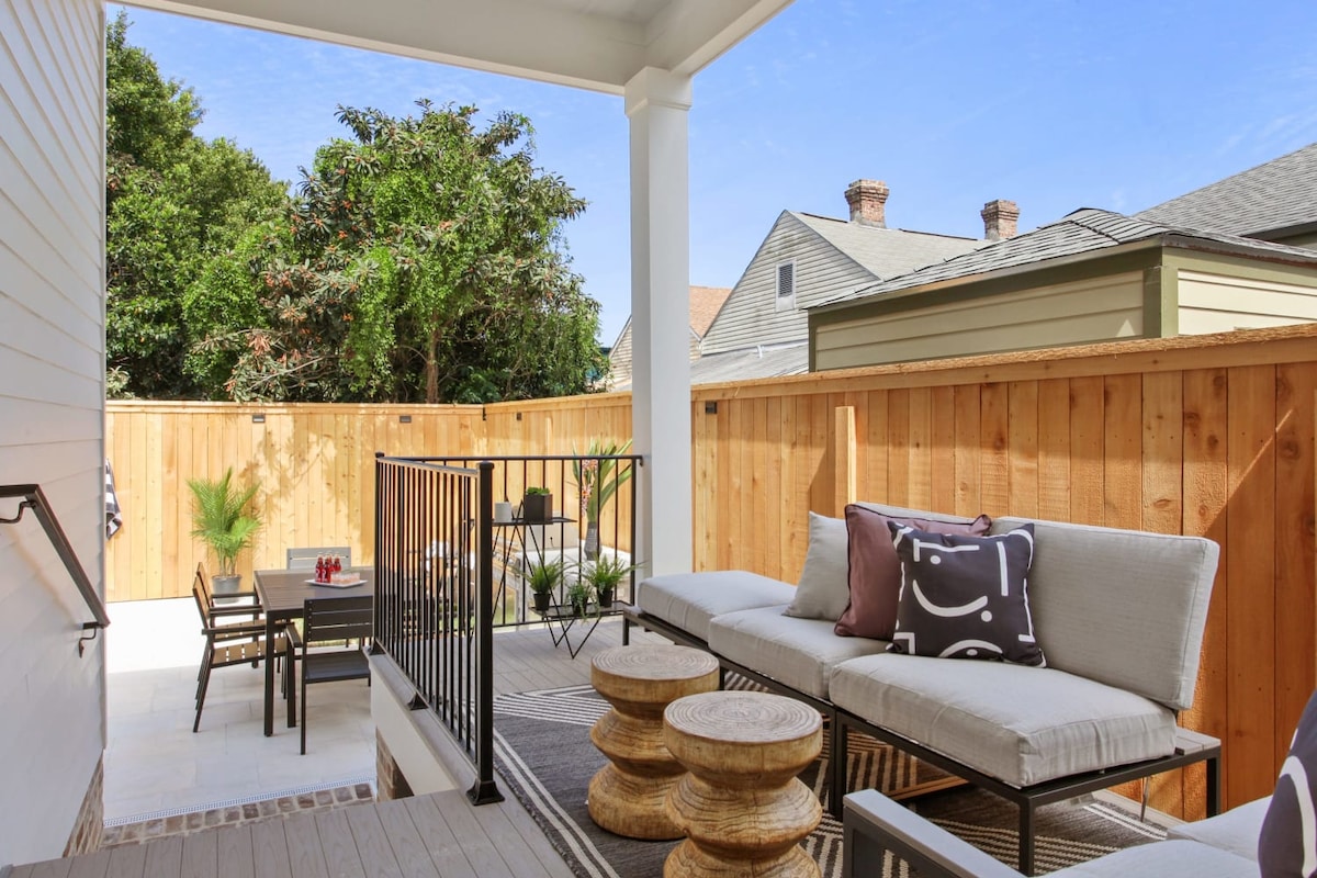 An outdoor patio area features comfortable seating with pillows and wooden accent tables. A dining set is positioned nearby under the shade of a roof, surrounded by wooden fencing and greenery, creating a relaxed environment for gatherings.