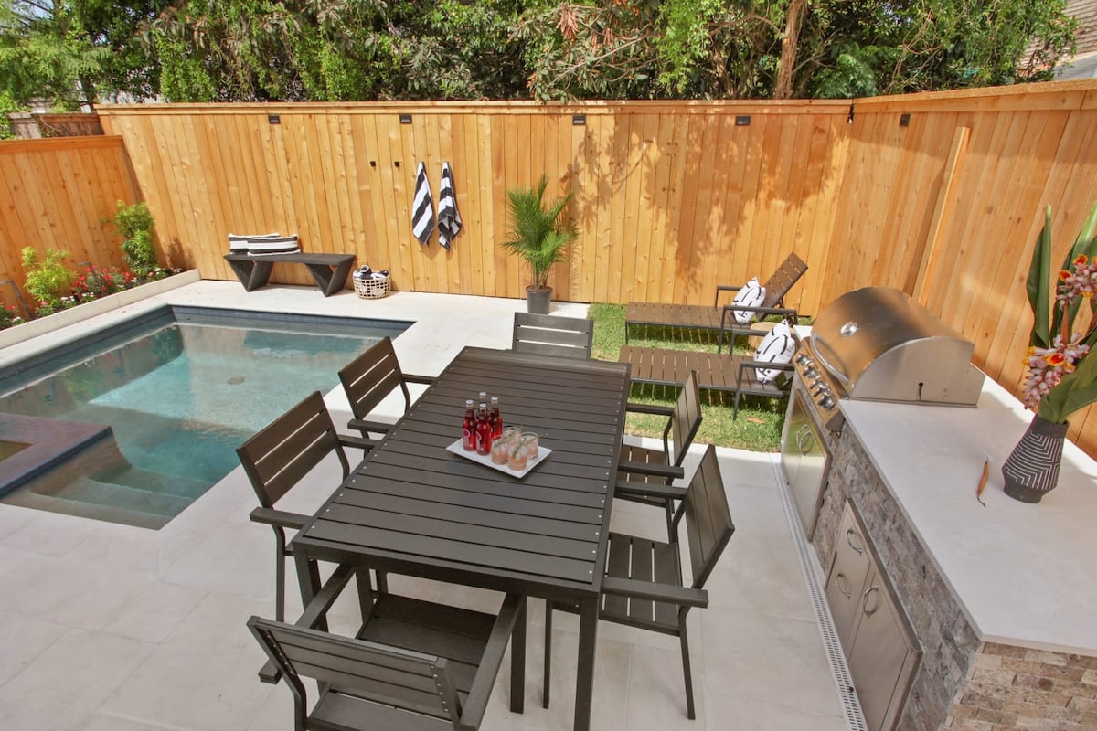 An outdoor space features a heated pool surrounded by a stone patio. A wooden dining table with eight chairs is set for dining. A grill is positioned nearby, with lounge chairs placed beside the pool. Lush greenery and a wooden fence provide privacy.