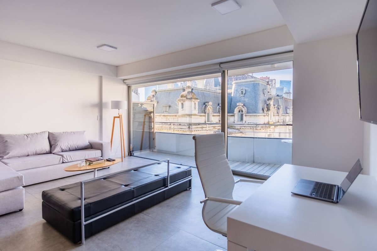 A spacious living area features large windows showcasing views of the city's architecture. A comfortable light-colored sofa is accompanied by a glass coffee table. A desk with a chair and a television mounted on the wall are also present, enhancing the functionality of the space.