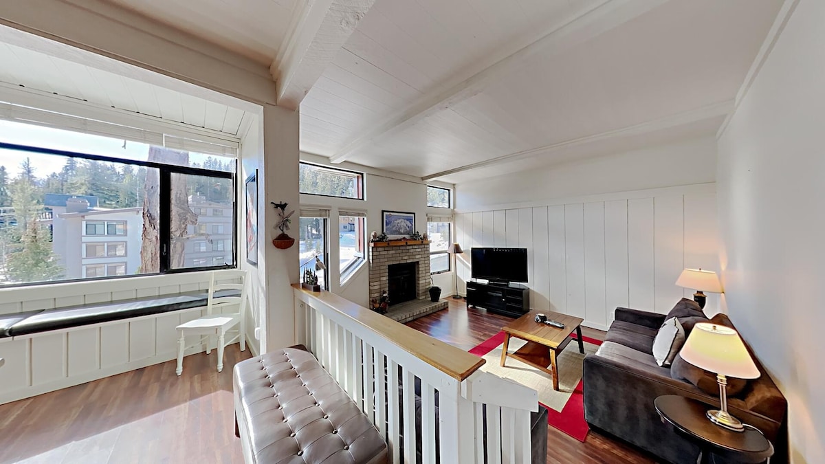 A well-lit living area is featured, showcasing large windows that invite natural light. Comfortable seating is provided on a dark sofa and a matching lounge chair. A coffee table is placed in front, with a fireplace nearby, enhancing the cozy ambiance of the space.