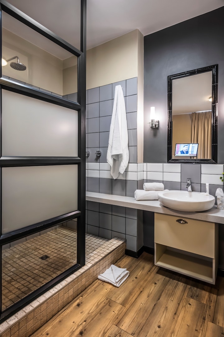 The bathroom is designed with a modern aesthetic. A shower area features a glass divider, and a large white basin is positioned on a stone countertop. Towels are neatly stacked on the counter, and wood-look flooring adds warmth to the space.