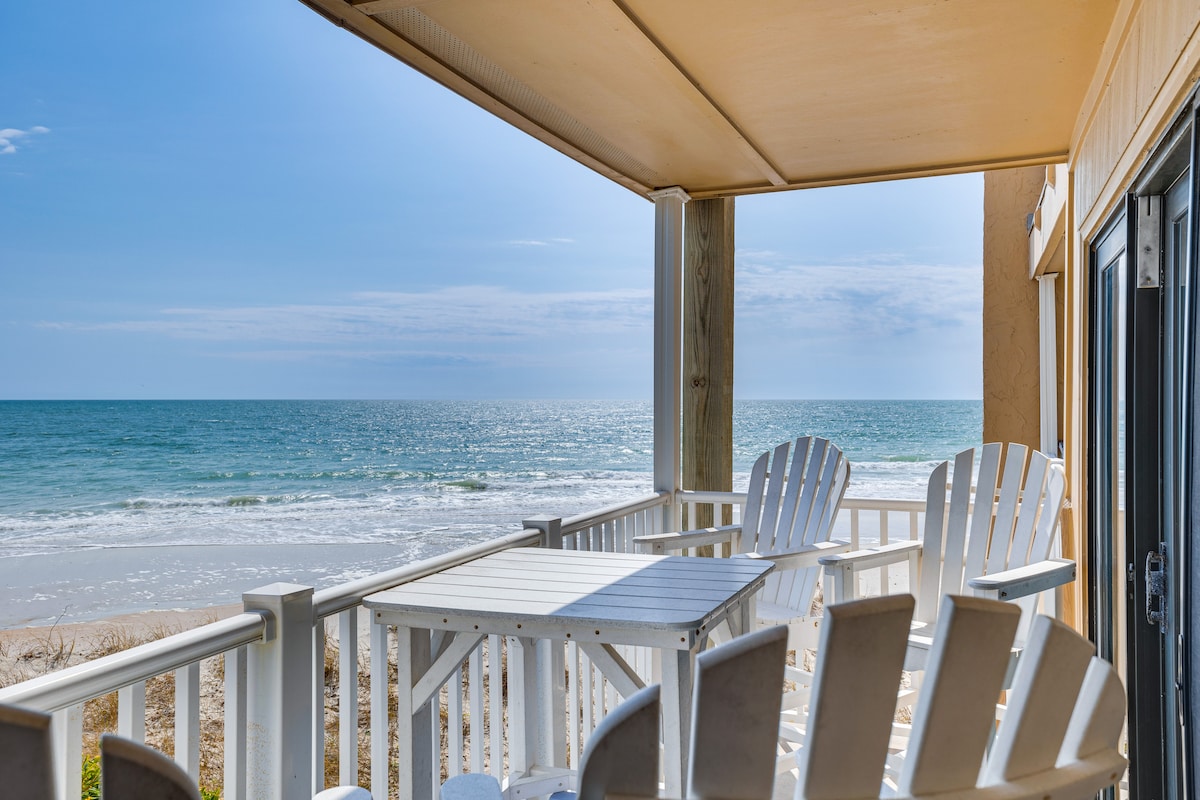 A furnished balcony overlooks the ocean, providing a tranquil setting with white adirondack chairs arranged around a white table. Sunlight reflects off the water, creating a serene atmosphere for relaxation and enjoyment of the coastal views.