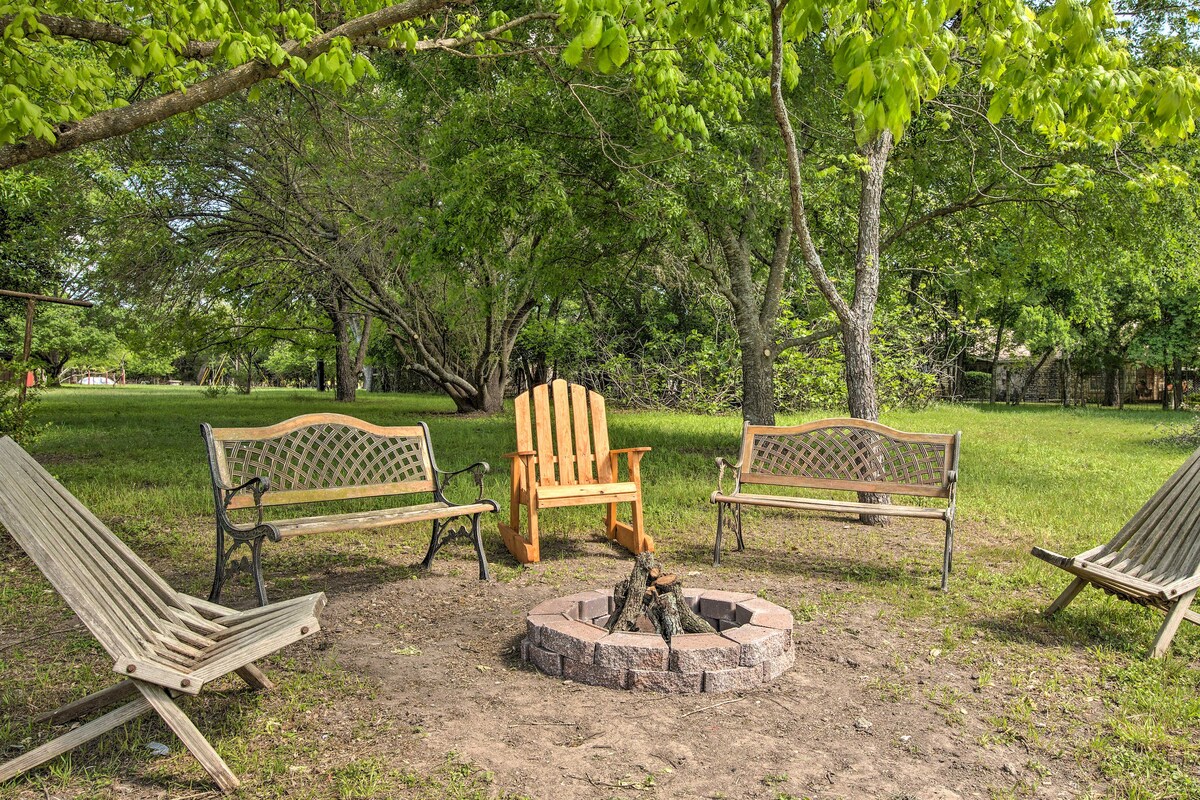 A fire pit is surrounded by several seating options, including two wooden adirondack chairs and two metal benches. The area is shaded by large trees, with a natural grassy landscape enhancing the outdoor experience.