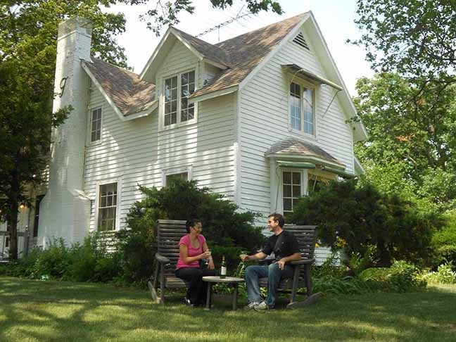 The white historic farmhouse is framed by lush greenery, featuring large windows and an inviting porch. Two individuals are seated on a bench, enjoying beverages amidst the serene outdoor setting.