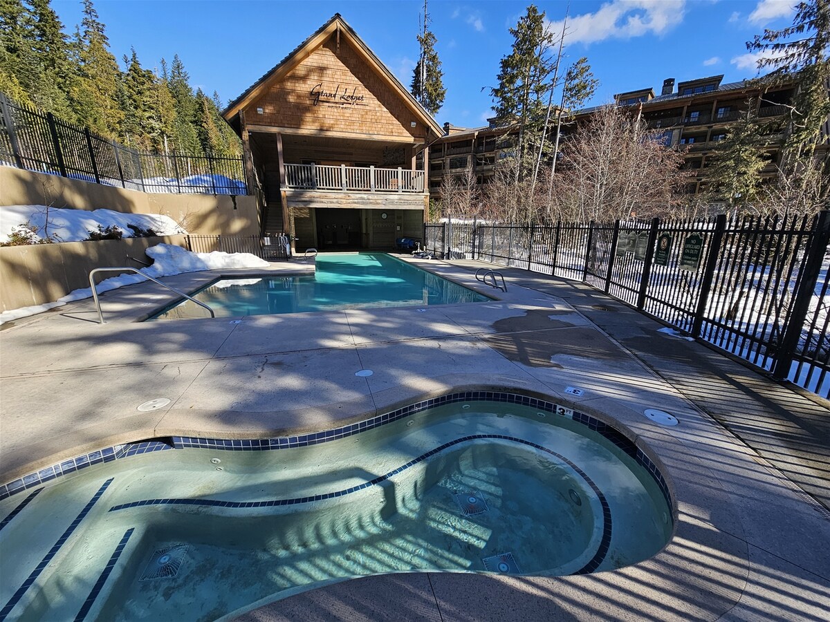 An outdoor heated pool and hot tub are set against a backdrop of tall trees and the Grand Lodge building. The pool area features a smooth concrete deck, surrounded by a decorative black fence, with snow visible in shaded areas reflecting a tranquil winter atmosphere.