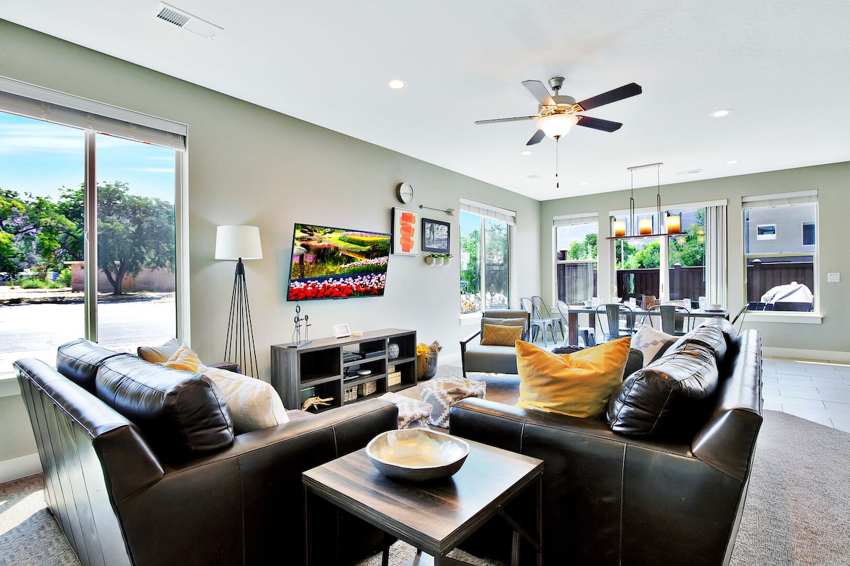 A spacious living area features a comfortable sectional sofa with decorative pillows, positioned around a low coffee table. A wall-mounted TV displays colorful artwork. Large windows let in natural light, showcasing views of landscaped grounds. A dining area with multiple chairs is visible nearby.