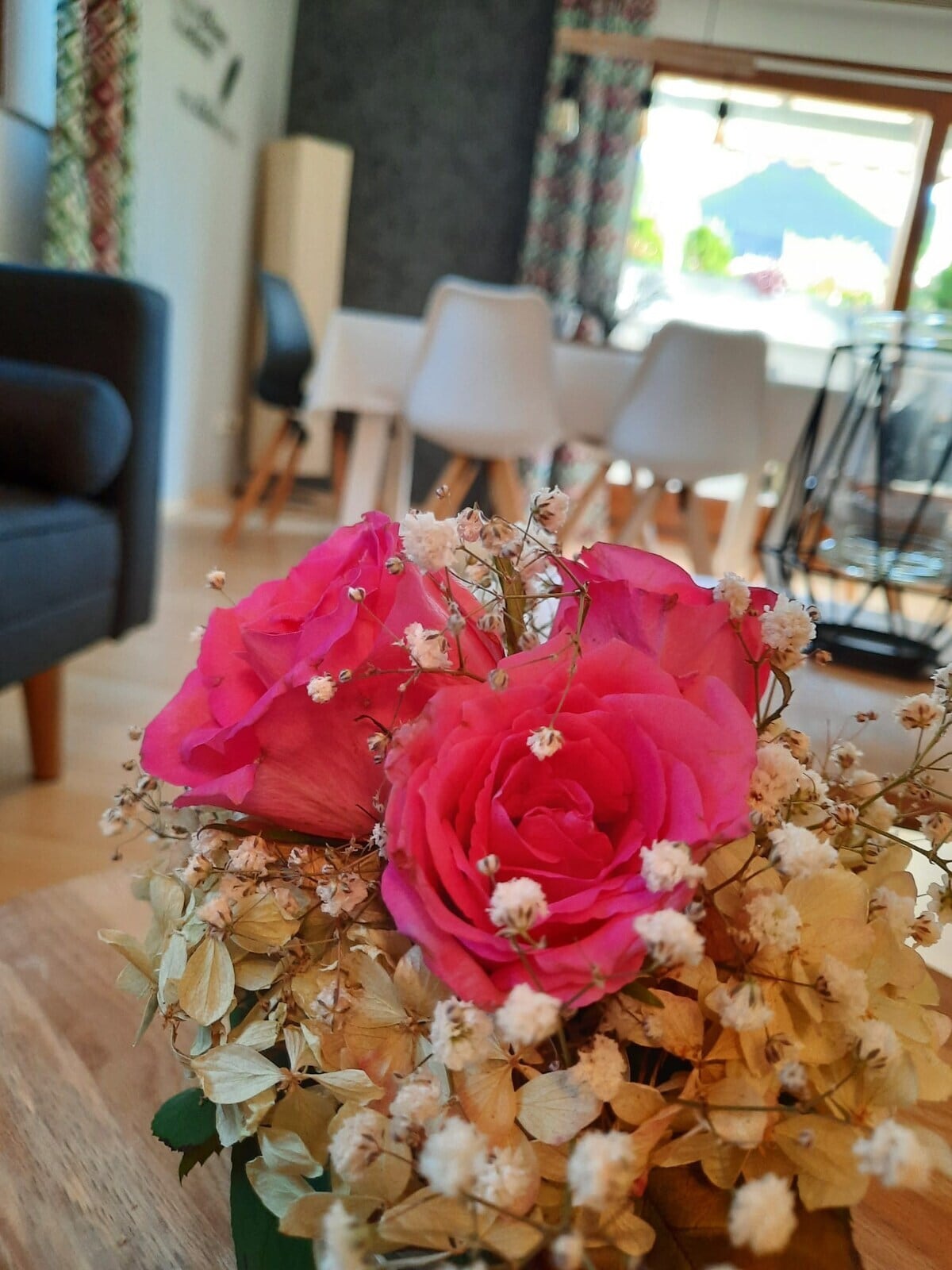 A floral arrangement featuring vibrant pink roses and delicate white filler flowers is positioned in the foreground. In the background, a dining area can be seen with a table and light-colored chairs, complemented by a decorative window view.