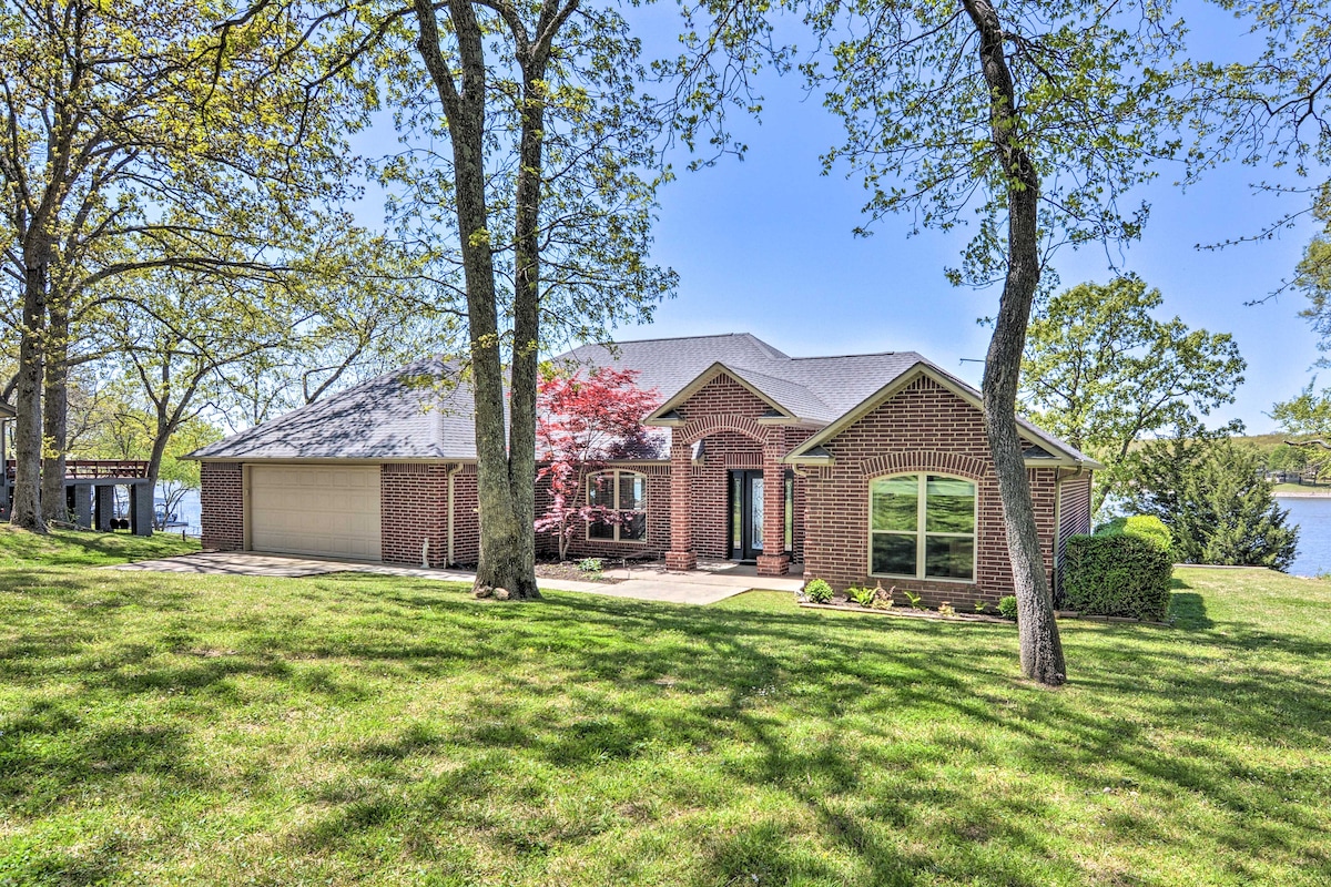 A brick exterior home is set amidst lush green grass and trees, with a scenic waterfront view visible in the background. Large windows reflect natural light, while the inviting pathway leads to the entrance, enhancing the property’s connection to nature.
