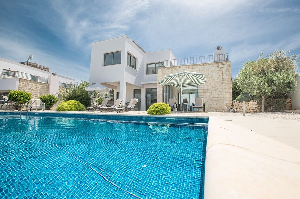 A view of the villa's exterior features a private pool framed by light-colored tiles. Lounge chairs are arranged nearby under a shaded area with an awning. The modern structure displays large windows and a balcony, set against a clear blue sky.