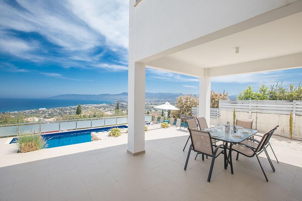 An outdoor dining area is featured under a covered terrace, equipped with a glass table and several chairs. The backdrop showcases scenic views of the sea and surrounding hills. A private pool is visible nearby, surrounded by sun loungers and greenery.