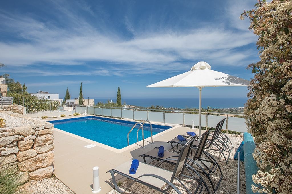 An outdoor pool area showcases clear blue water, flanked by sun loungers and a large umbrella. Lush greenery and flowering plants add a natural touch, while distant views of the sea and sky can be seen beyond the glass railing.