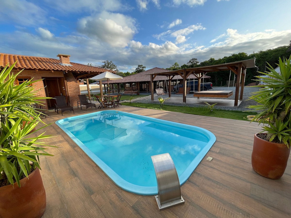 A refreshing swimming pool is surrounded by wooden decking and potted green plants. The outdoor area features shaded seating and dining spaces. In the background, rustic structures and an expansive lawn create a welcoming atmosphere, complemented by the soft light of the late afternoon.