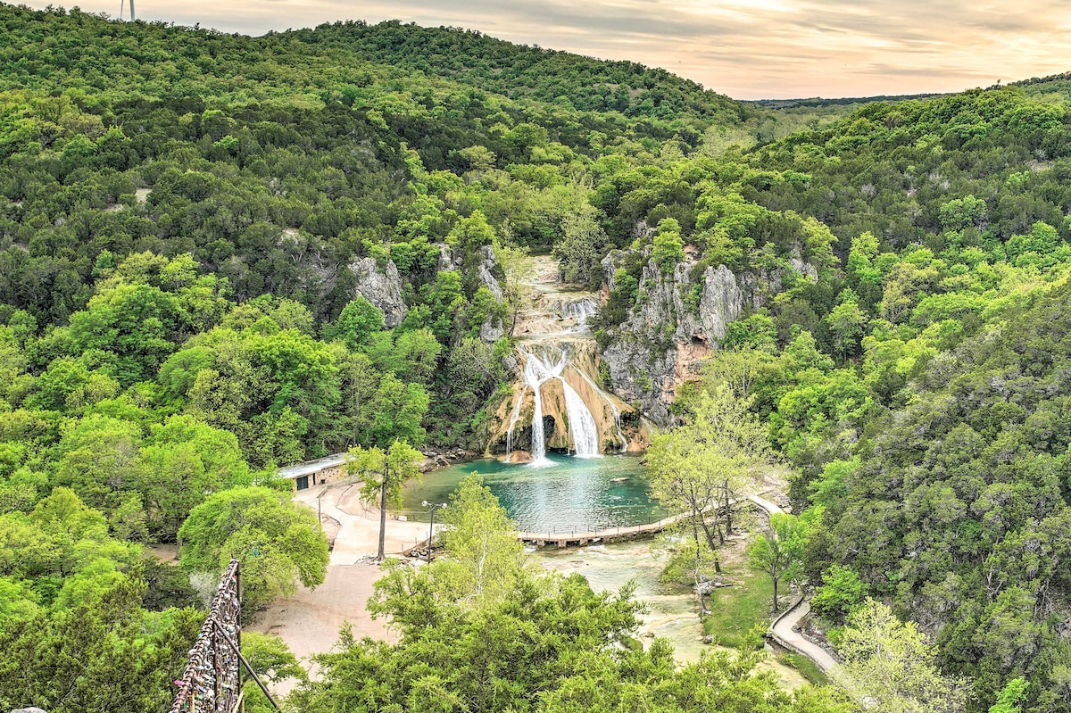 A stunning view showcases the 77-foot waterfall cascading into a serene pool, surrounded by lush greenery. The vibrant trees frame the scene, highlighting the natural beauty of the Arbuckle Mountains in the soft light of dusk.