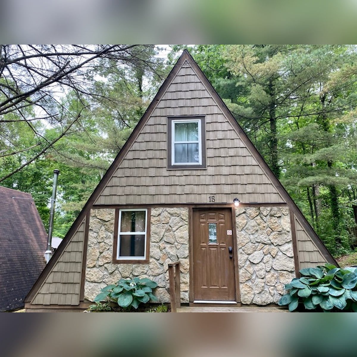 An A-Frame cabin is showcased, featuring a peaked roof and a stone facade at the base. A single window is positioned above the door, framed by wood siding. Lush greenery surrounds the exterior, contributing to a secluded, woodland appearance.