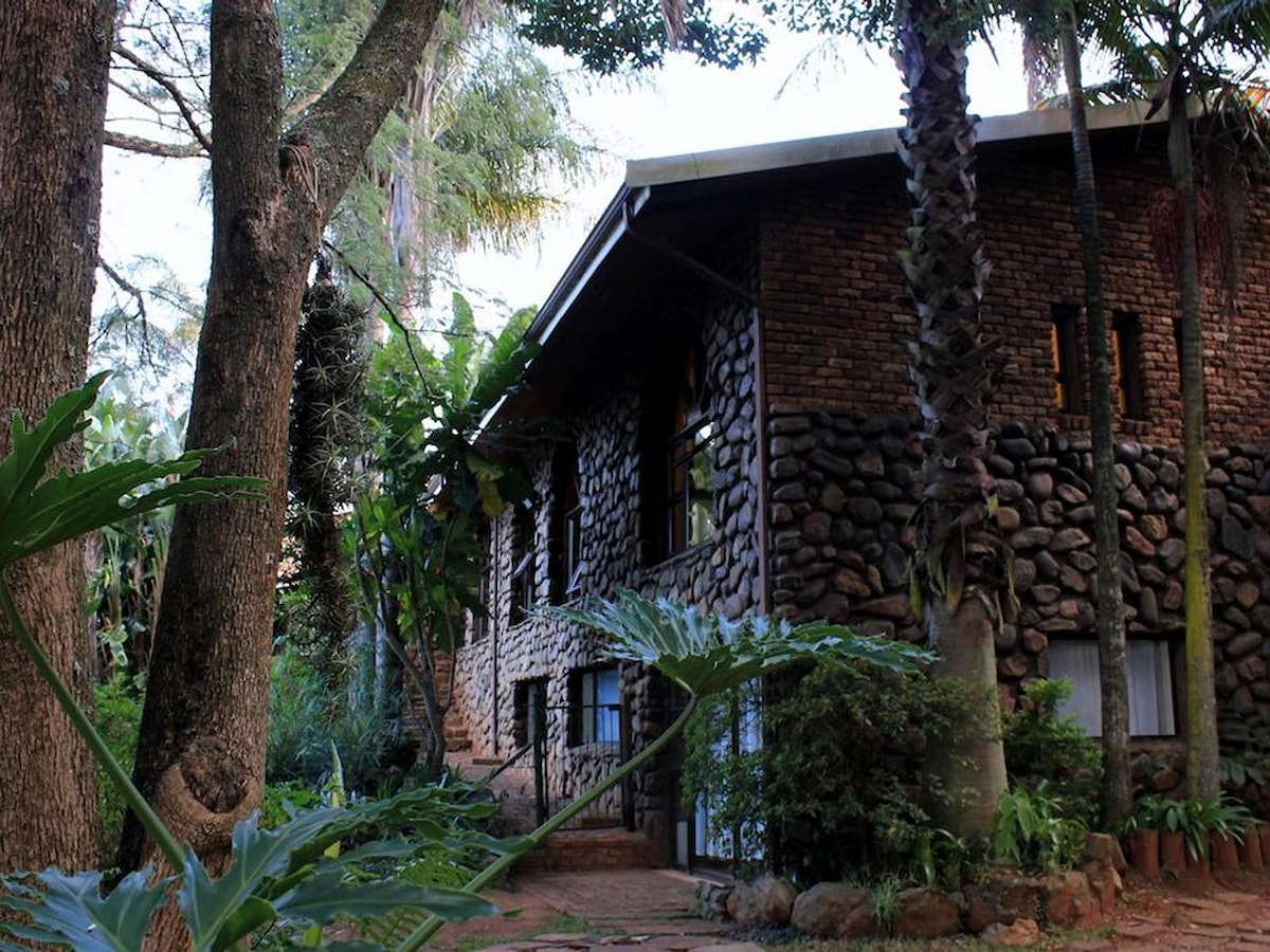 A rustic stone building is surrounded by lush greenery and tall trees. Large windows allow natural light to enter the spaces inside. The natural landscape includes various plants, contributing to a serene and inviting environment.