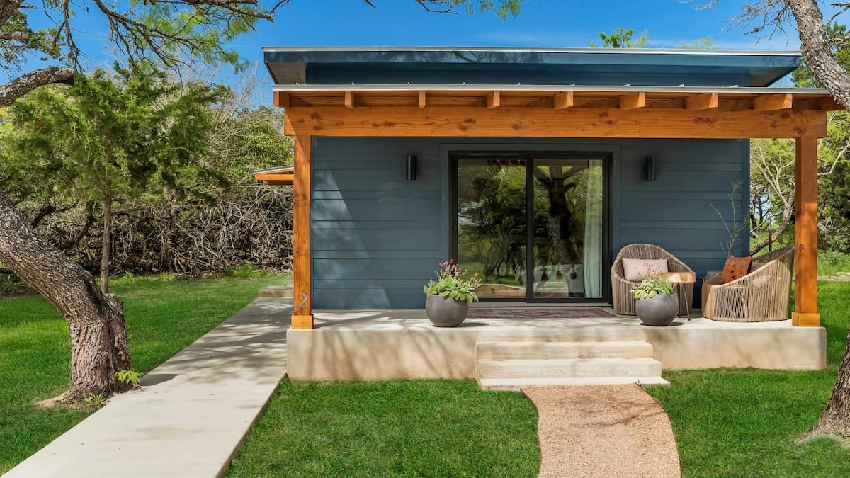 A modern cabin features a welcoming entrance with wooden beams above a raised porch. Two stylish chairs are positioned beside potted plants, while a stone pathway leads from the porch through the well-maintained green lawn, surrounded by natural landscapes.