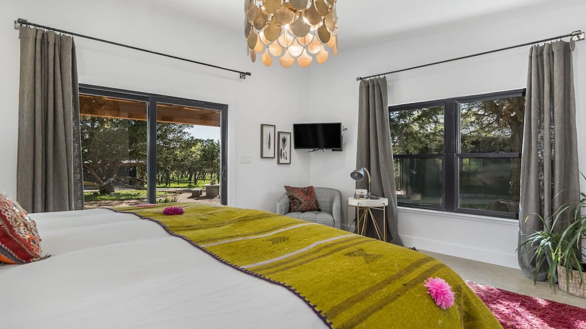 A spacious bedroom features large windows framing views of the surrounding nature. A plush bed is dressed in soft linens and a vibrant blanket, adding a pop of color. A small television is mounted on the wall, and a cozy seating area is visible beside a decorative plant.