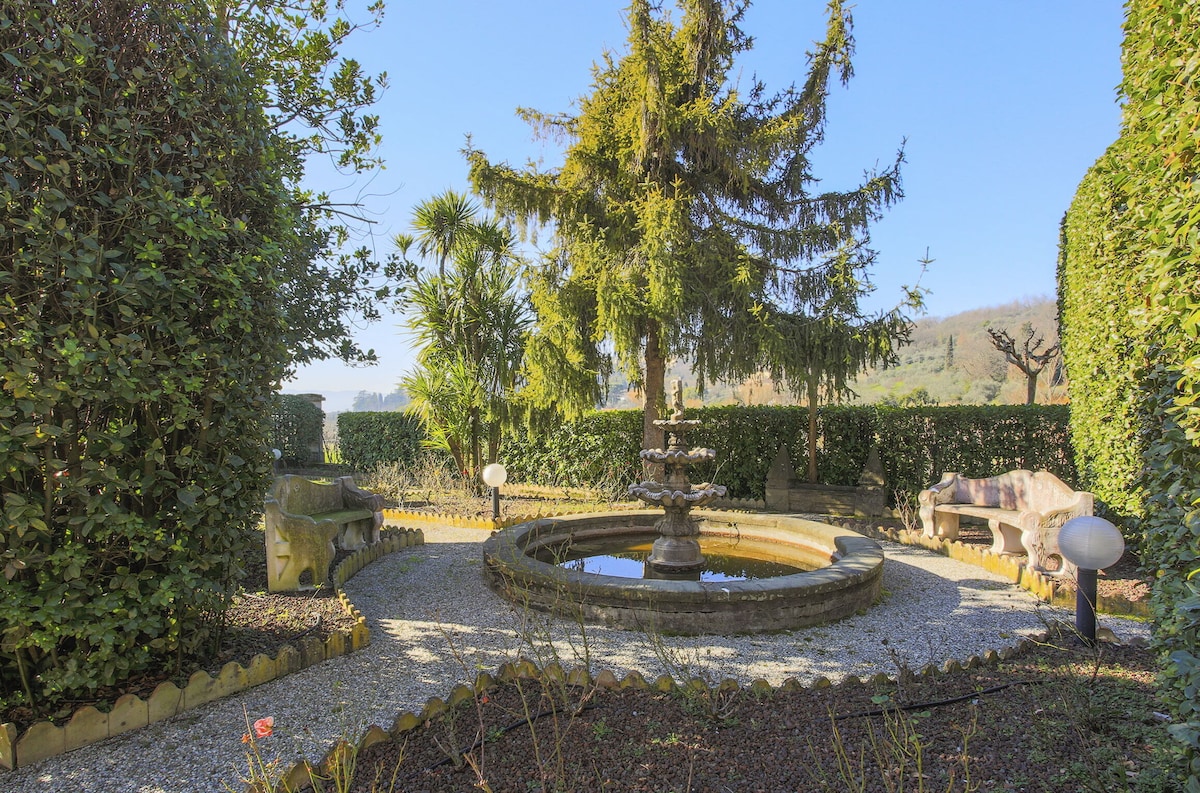 A serene garden features a circular fountain at its center, surrounded by shrubs and trees. Stone benches are placed around the fountain, providing a peaceful setting. Gravel pathways lead through landscaped greenery, creating a tranquil atmosphere for relaxation.