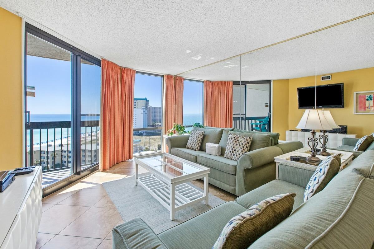 The spacious living area features large windows that provide stunning views of the Gulf of Mexico, allowing natural light to brighten the room. Comfortable seating, including a sectional sofa and accent chairs, is arranged around a glass coffee table, creating a welcoming space for relaxation.
