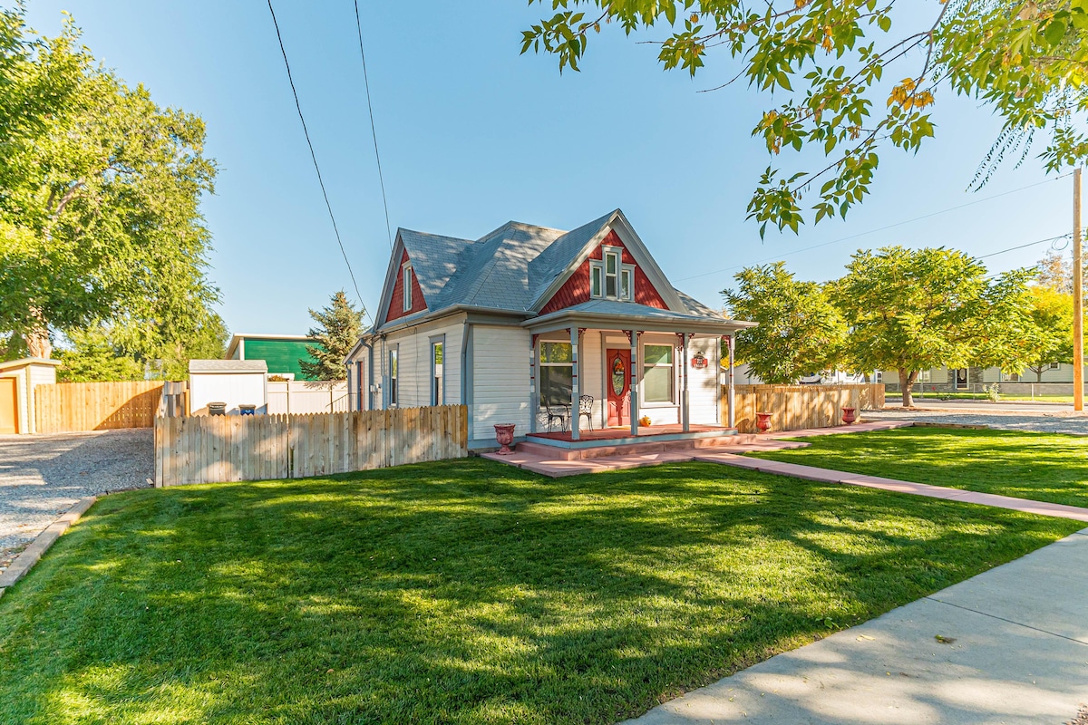A charming two-story house features a well-maintained lawn framed by neatly trimmed trees. The inviting porch, accented with red details, provides seating for outdoor relaxation. A gravel driveway leads to additional outdoor space, enhancing the welcoming curb appeal of the property.