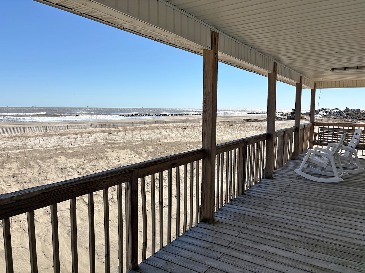 A spacious wooden deck overlooks the beach and ocean, with white rocking chairs positioned for relaxation. The sandy beach extends toward the water's edge, while gentle waves can be seen lapping against the shore under a clear blue sky.