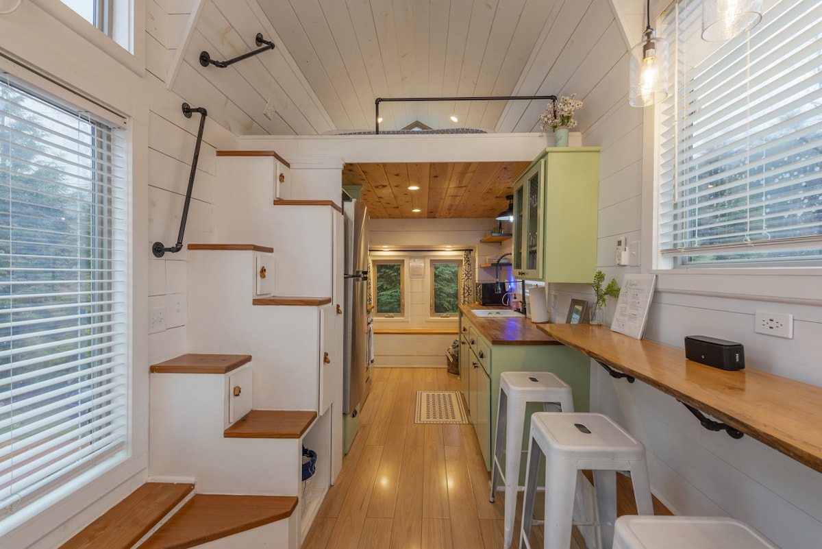 The interior features a compact kitchen with sleek green cabinetry and open shelving. A wooden countertop offers space for meal preparation. Stairs lead to a lofted sleeping area, while large windows provide ample light and views of the surrounding greenery.