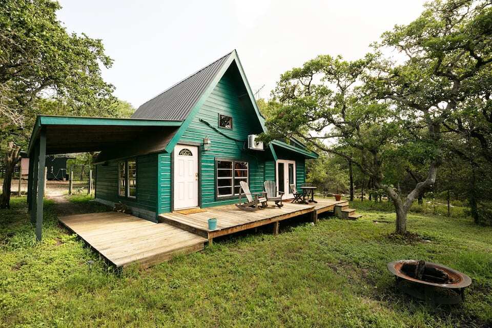 A vibrant green A-frame cabin is nestled among lush trees, featuring a wide wooden deck with seating. The entrance is adorned with dual doors, inviting guests to explore the serene surroundings. A rustic firepit is situated in the grassy area nearby, enhancing the outdoor experience.