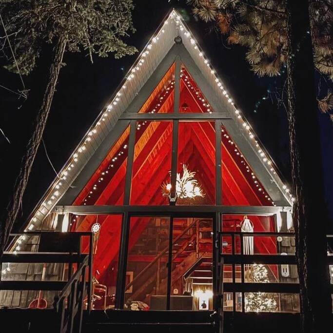 The A-frame cabin features a striking triangular silhouette, illuminated by string lights. A large glass wall allows views of the inviting interior, showcasing wood accents and a high ceiling. The surrounding trees add to the serene ambiance of this mountain retreat.
