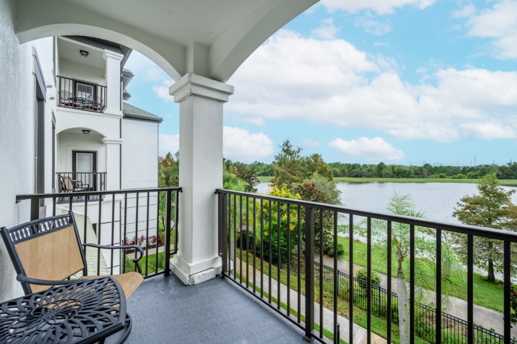A private balcony features two chairs and a round table, offering scenic views of a serene lake and lush greenery. The architecture includes elegant columns and a spacious overhang, enhancing the welcoming outdoor space.