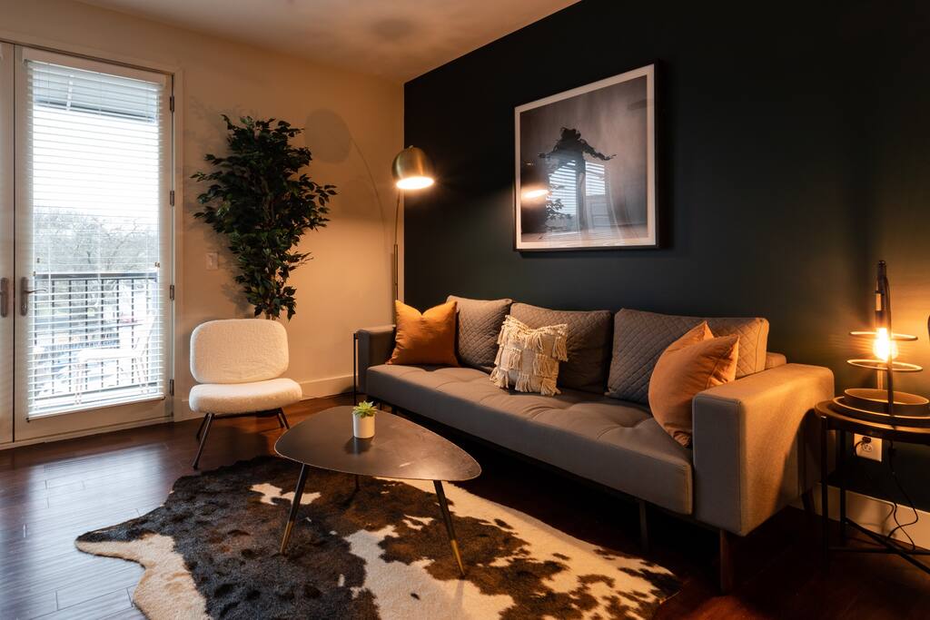 A modern living area is highlighted by a plush gray sofa adorned with decorative pillows, positioned beside a sleek coffee table. A floor lamp provides warm lighting, while a potted plant adds a touch of greenery. Large windows allow natural light to fill the space.