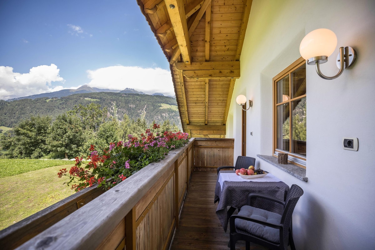 A balcony featuring a wooden railing adorned with colorful flowers offers a scenic view of the surrounding mountains and valley. A small table with fruit sits beside two chair, providing a serene space to relax outdoors.