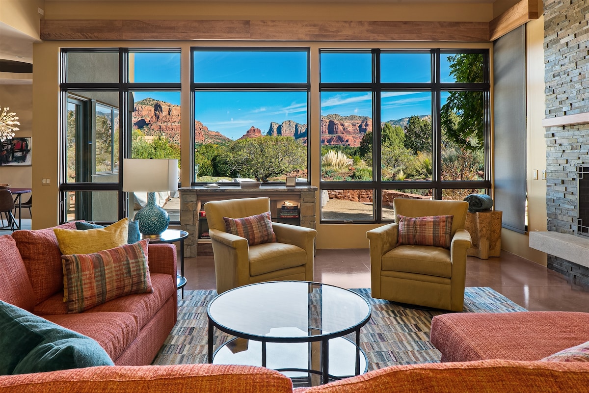 A spacious living area features large picture windows that offer expansive views of the Sedona peaks. Comfortable seating is arranged around a round glass table, complemented by a cozy fireplace and eclectic decor. Earthy tones and colorful accents create a welcoming environment.