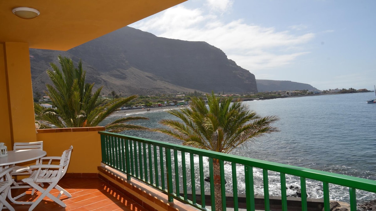 The terrace features a green railing overlooking the sea and nearby mountains. Sunlight reflects on the water, while palm trees add a tropical touch. Dining chairs and a table are arranged to take in the views.