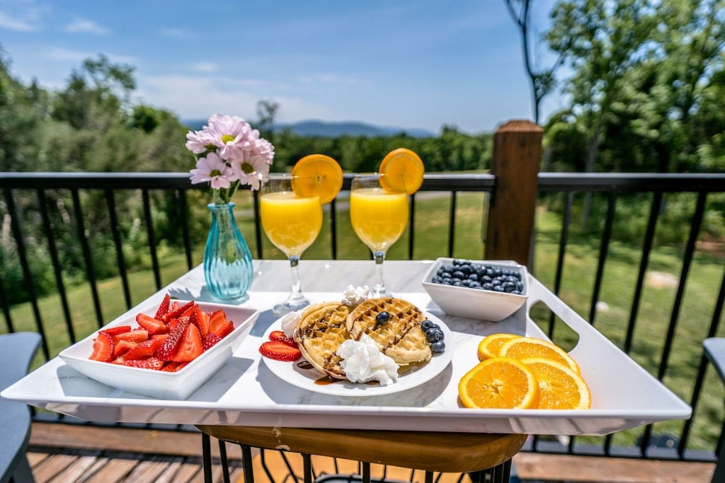 Breakfast is presented on a white tray featuring pancakes topped with whipped cream and blueberries, surrounded by fresh strawberries and orange slices. Two glasses of orange juice are adorned with orange slices, and a small vase holds pink flowers, creating a welcoming breakfast setting with a scenic view.