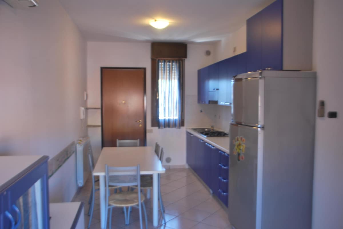 A compact kitchenette features blue cabinetry and stainless steel appliances, including a refrigerator and stove. A small dining table with four chairs is positioned beside the entrance. Natural light enters through a window dressed with patterned curtains, illuminating the tiled floor.