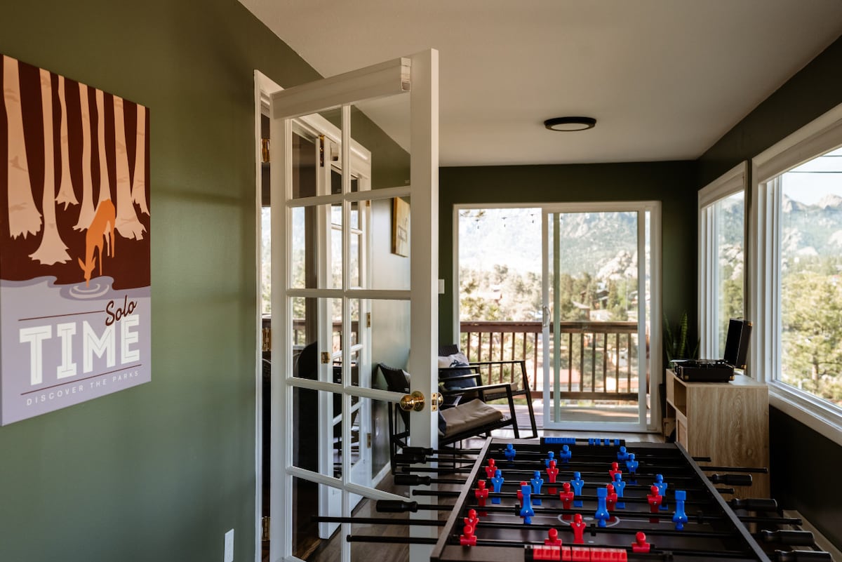 A game room is depicted with a foosball table in the foreground, featuring red and blue players. French doors open to a scenic view of the mountains, while a cozy chair and a poster on the green wall add character to the playful space.
