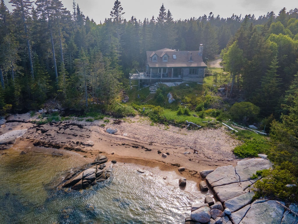The log cabin is positioned on a sandy beach, surrounded by trees and rocky shoreline. The gentle waves lap at the water's edge, while the two-story structure features a sloped roof and a stone chimney. Natural elements create a serene, secluded atmosphere.