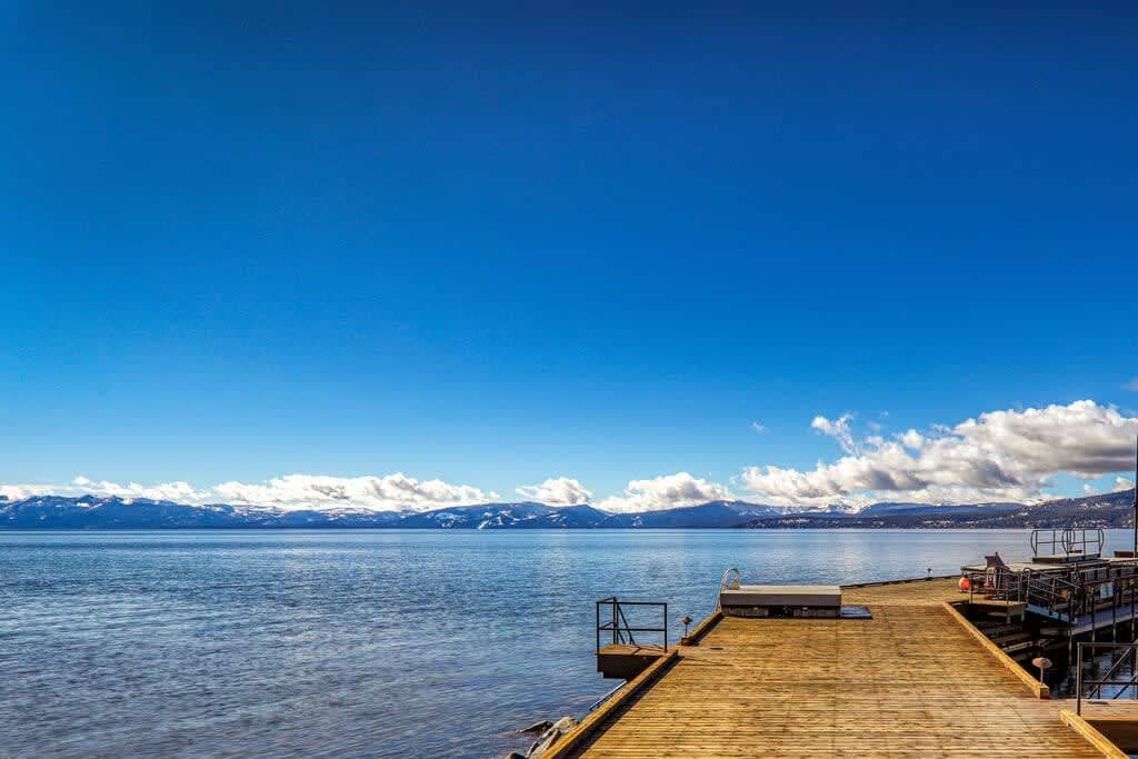 A tranquil lakeside view is captured, featuring a wooden pier extending into calm waters. The expansive blue sky above is dotted with fluffy white clouds, while distant mountains line the horizon, creating a serene backdrop.