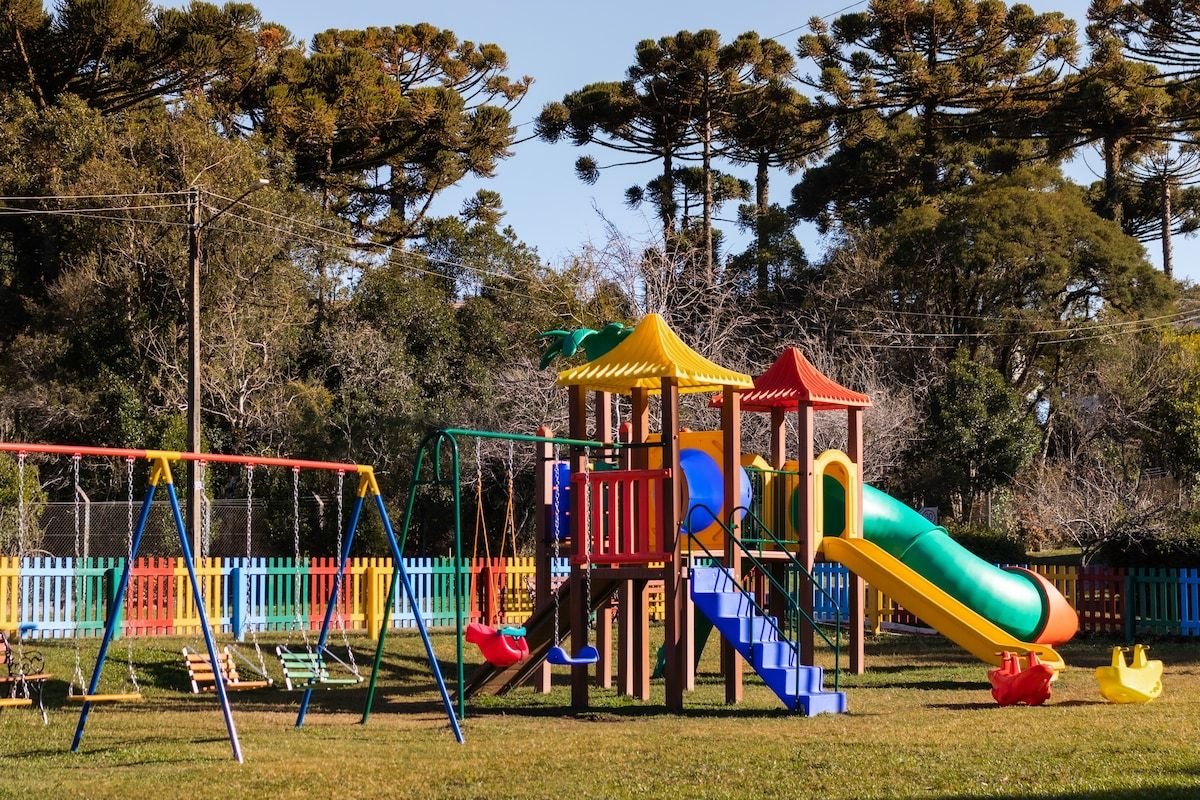 A vibrant playground is set within a grassy area, featuring colorful slides, swings, and play structures. The equipment includes a climbing frame with bright roofs and a slide. Surrounding trees provide a natural backdrop, while a colorful picket fence encloses the space.