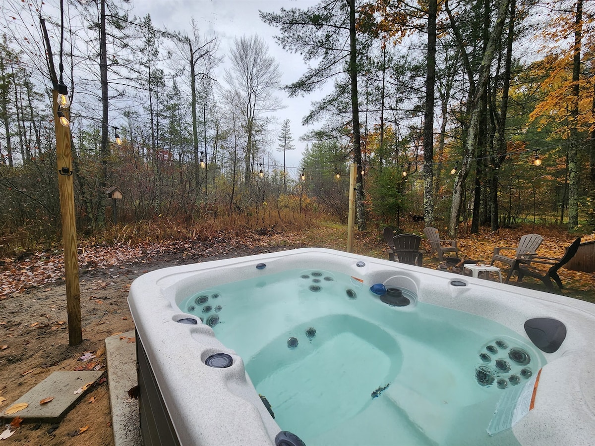 A spacious hot tub is situated on a sandy area surrounded by trees, offering a secluded retreat. The clear water is framed by warm lighting and nearby seating, inviting relaxation amidst a natural landscape. The setting features autumn foliage, enhancing the tranquil atmosphere.