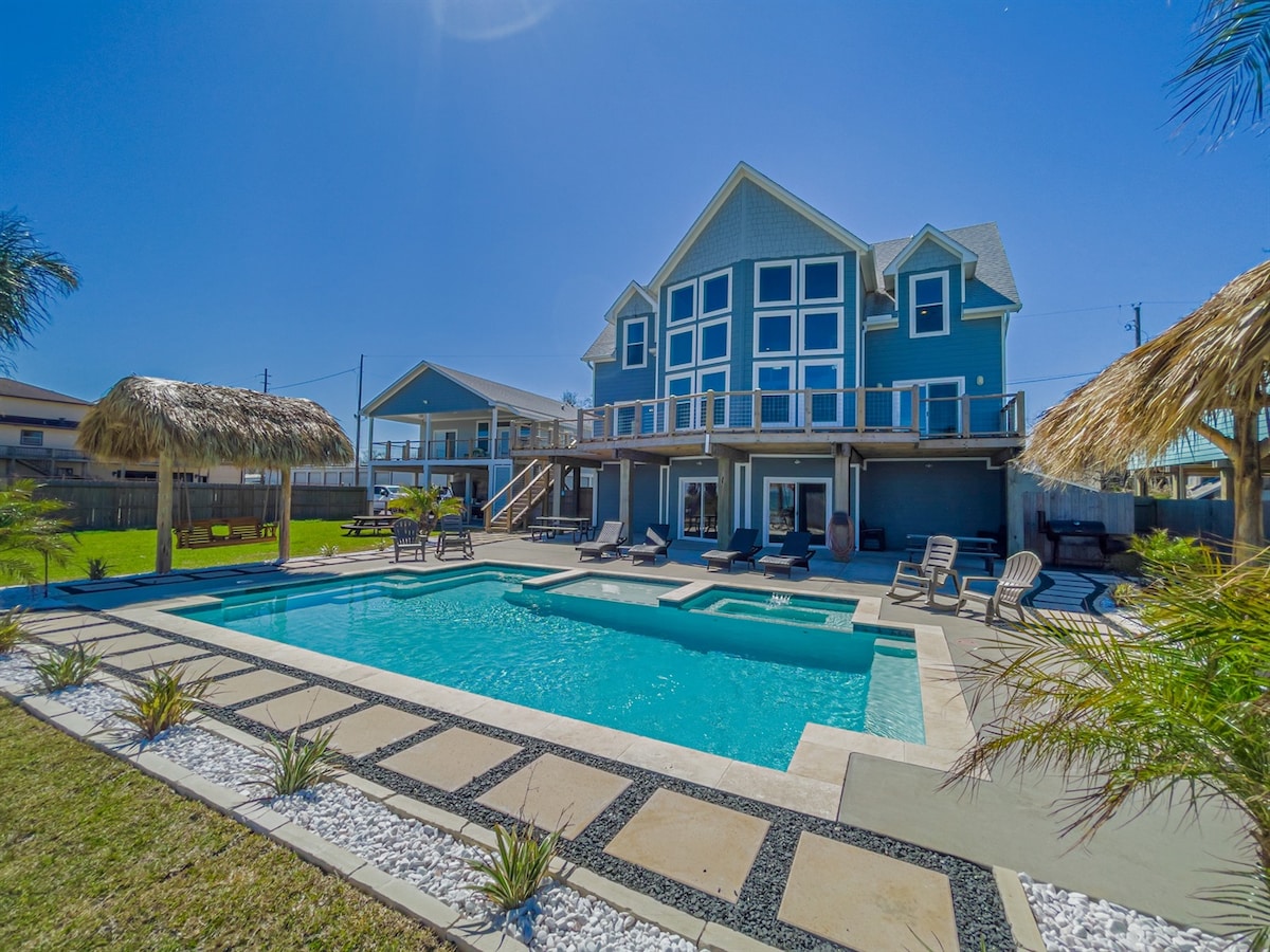 A spacious outdoor area is presented with a crystal-clear pool surrounded by lounge chairs and tropical landscaping. The two-story lodge is showcased in the background with multiple windows, while palapas provide shaded seating. The bright blue sky creates a vibrant atmosphere.