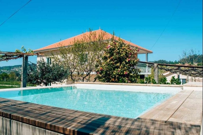 A modern house with a red tiled roof is set against a clear blue sky. In the foreground, a small pool is surrounded by a wooden deck and well-maintained greenery, including trees and flowering plants, creating a serene outdoor setting.