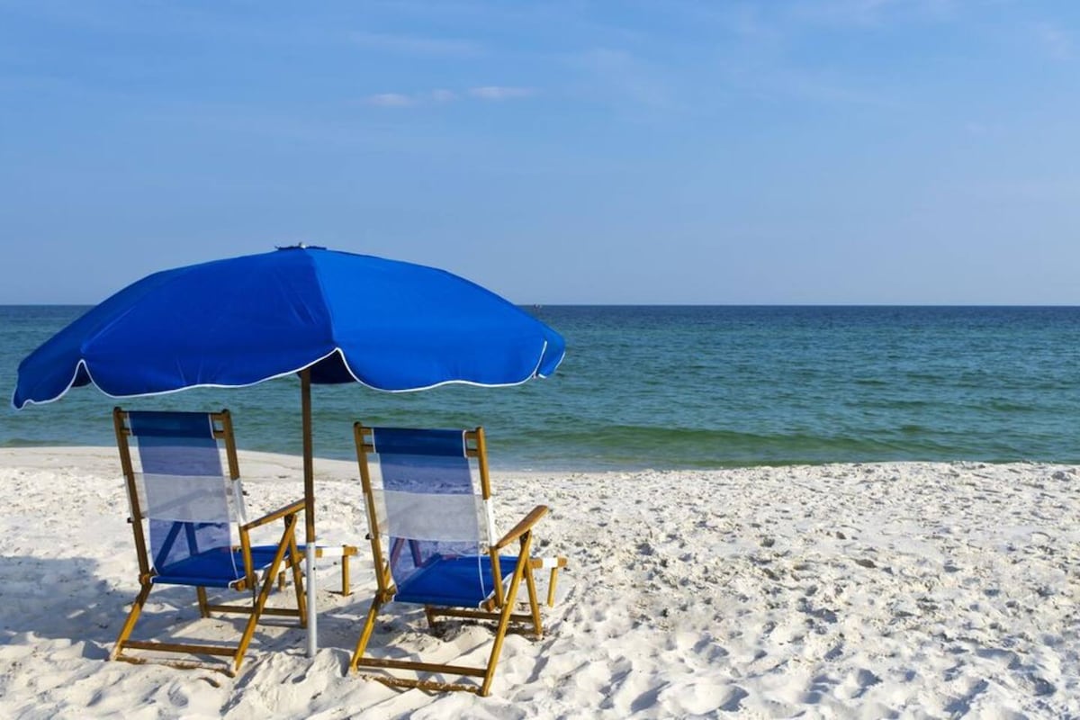 Two beach chairs are positioned beneath a large blue umbrella on a sandy beach. The calm turquoise water extends to the horizon, with gentle waves lapping at the shore. The bright sky complements the serene coastal scene.