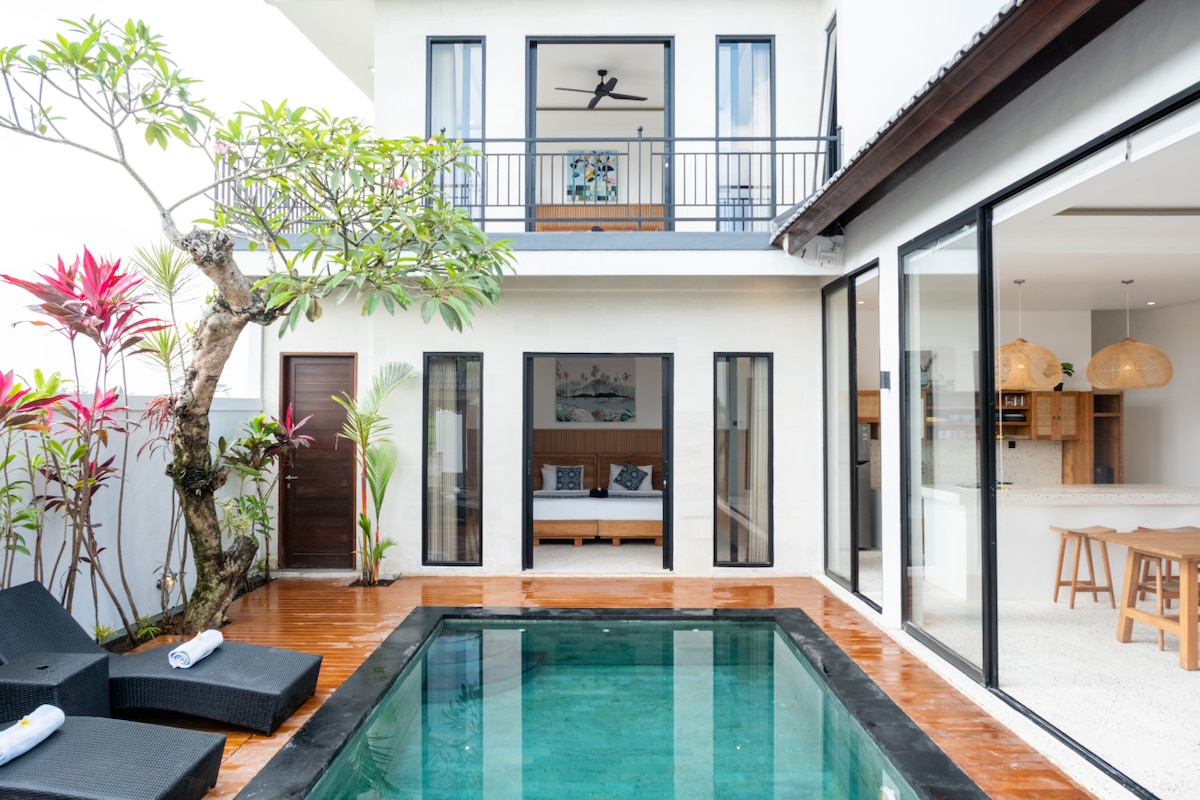 A private swimming pool is framed by stylish lounge chairs. The modern villa's two-story design is highlighted by large glass doors and windows, providing views of the lush garden and inviting living area. Natural light fills the open space, creating a bright and airy atmosphere.