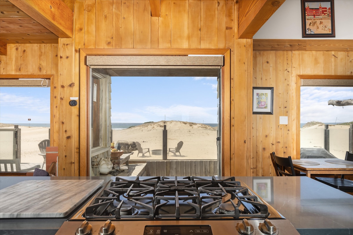 The image captures a view from inside the home, looking through large glass doors that reveal a sandy beach and ocean. Warm wooden walls and exposed beams add a rustic touch. The kitchen countertop is visible in the foreground, complemented by a gas stovetop.