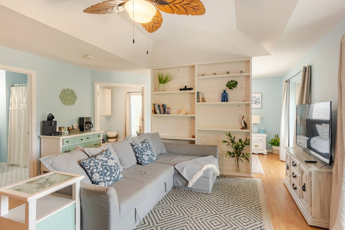 A spacious living area features a light blue color palette and tasteful coastal decor. A large gray couch adorned with patterned throw pillows is positioned in front of an entertainment unit. Natural light filters through the nearby window, enhancing the open layout.