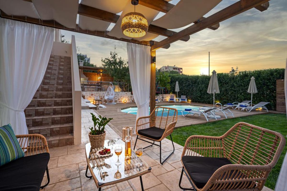 An outdoor seating area features comfortable chairs around a glass table, adorned with drinks and snacks. Soft drapes provide privacy, while a swimming pool is visible in the background. Umbrellas and sunbeds are arranged invitingly in the well-maintained garden, bathed in warm sunset light.